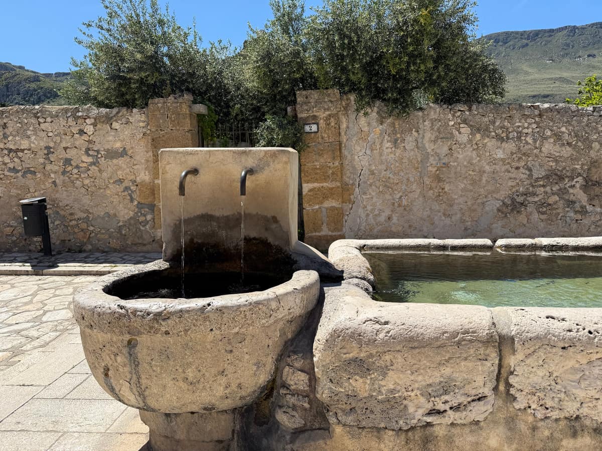 Fontana in pietra con due getti d’acqua e una lunga vasca, davanti a un muro a secco e a una siepe di ulivi