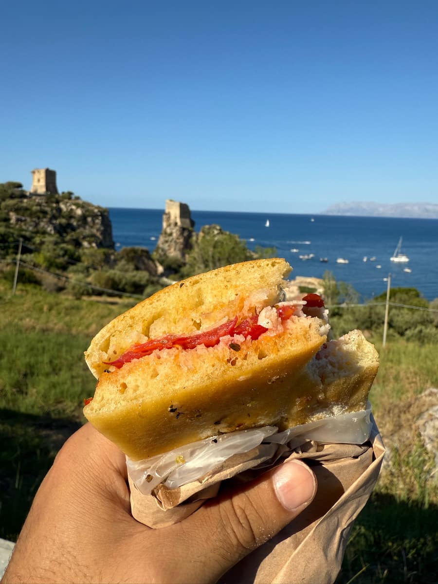 Una mano tiene un pane cunzato con pomodoro e formaggio; sullo sfondo le torri della Tonnara, i faraglioni e il mare blu con barche a vela