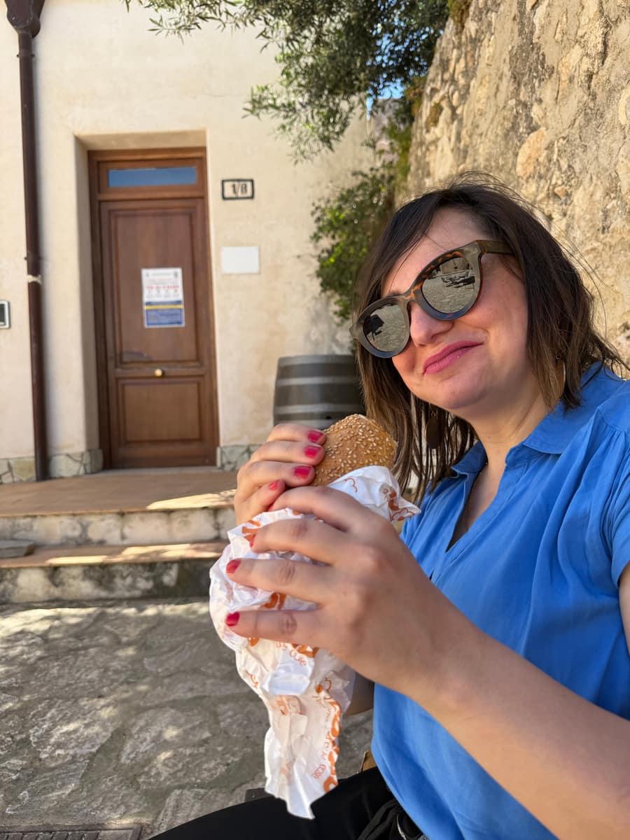 Una donna con gli occhiali da sole sorride mentre tiene un panino al sesamo appena comprato, seduta davanti a una casa del borgo
