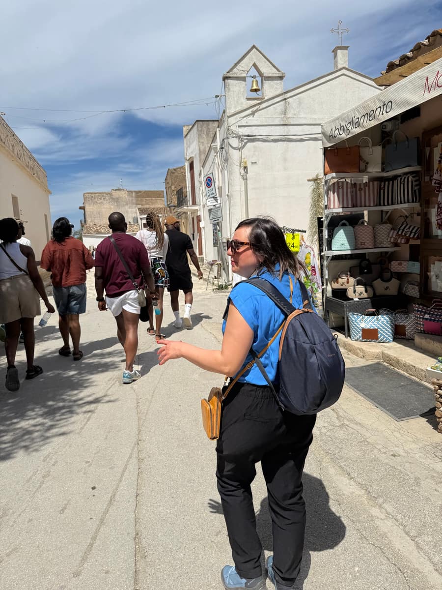 Una donna con lo zainetto cammina nella via del borgo, accanto a un negozio di borse e a una chiesetta bianca con la campana