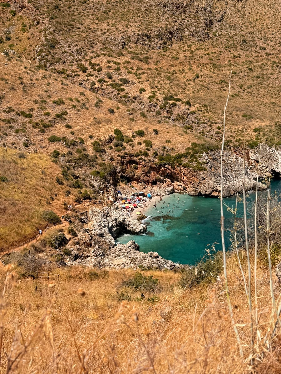 Cala Capreria vista dall’alto: spiaggia di ciottoli, ombrelloni e acqua turchese tra i pendii della Riserva